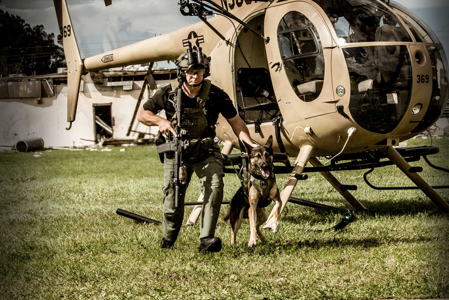 Police officer in tactical gear with service dog near helicopter on grassy field.